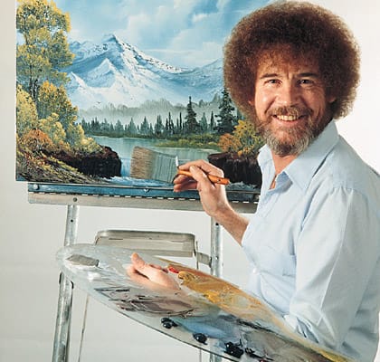 Bob Ross paints a misty mountain and lake scene on set of The Joy of Painting, his brush hovering as he adds a stand of evergreens.