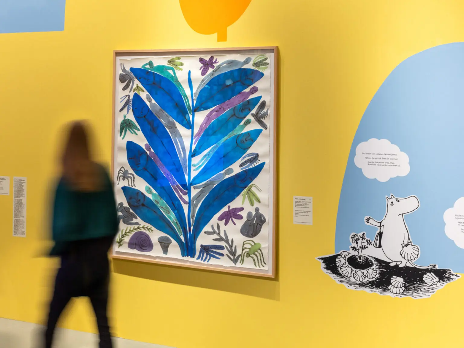 A visitor walking through the Escape to Moominvalley exhibition at the Architecture & Design Museum, with illustrated artworks.