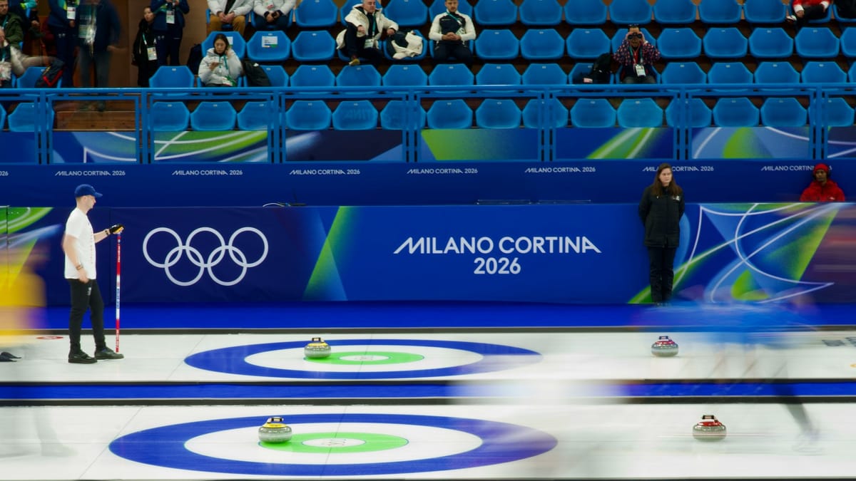 The curling venue during the Milano Cortina 2026 Winter Olympics, with athletes competing on the ice under arena lighting.