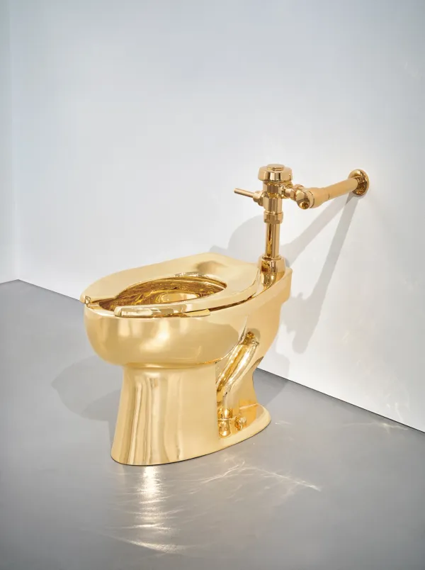 Gleaming 18-karat gold toilet in a stark white gallery, its curved surfaces reflecting spotlights like liquid metal.